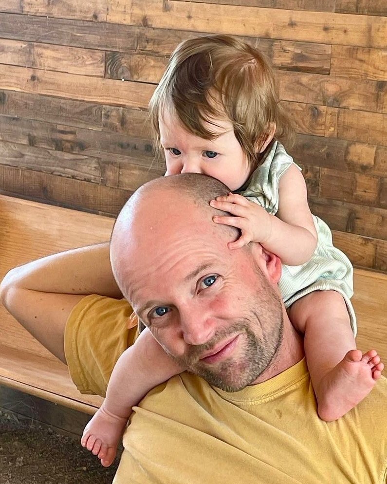 A Saltwater partner smiling, with a toddler riding on their shoulders, in front of a weathered wood wall